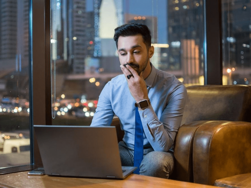 A man in a business casual outfit sitting in an armchair, looking at a laptop screen with a thoughtful expression, with city lights visible through the window in the background.