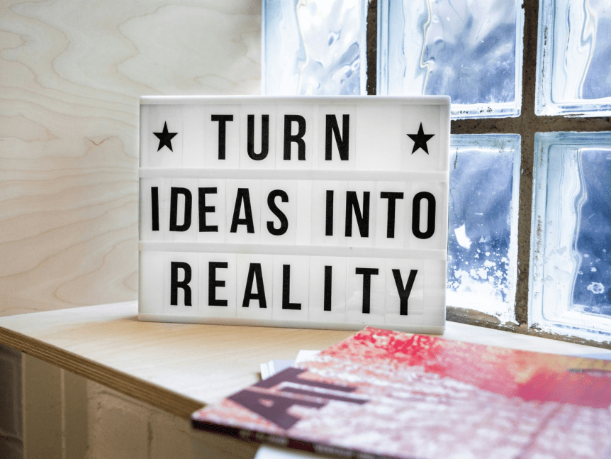 A lightbox with the phrase "TURN IDEAS INTO REALITY" in black letters, placed on a shelf near a glass block window, with magazines partially visible in the foreground.